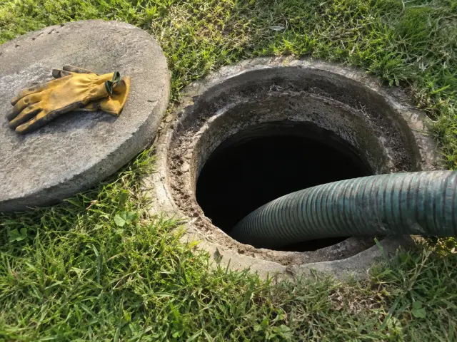 Manguera de succión entrando en la cámara abierta de una fosa séptica, con guantes sobre la tapa